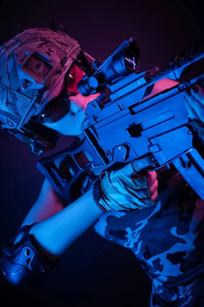 A menina em macacão militar airsoft posando com uma arma em suas mãos em um fundo escuro na névoa em luz de néon — Fotografia de Stock