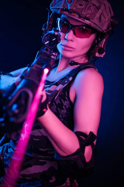 Dívka ve vojenských kombinézách airsoft pózuje s pistolí v rukou na tmavém pozadí v oparu v neonovém světle — Stock fotografie