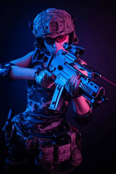 La chica en mono militar airsoft posando con una pistola en sus manos sobre un fondo oscuro en la neblina en luz de neón — Foto de Stock