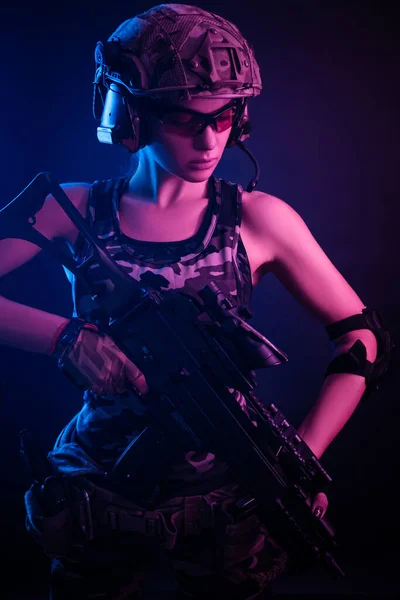 Девушка в военном комбинезоне аэрозоль позирует с пистолетом в руках на темном фоне в тумане в неоновом свете — стоковое фото