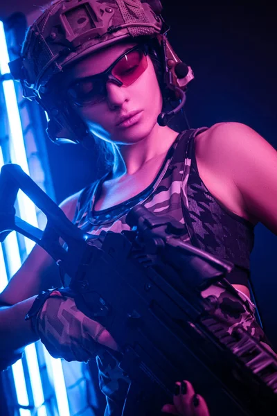 A menina em macacão militar airsoft posando com uma arma em suas mãos em um fundo escuro na névoa em luz de néon — Fotografia de Stock