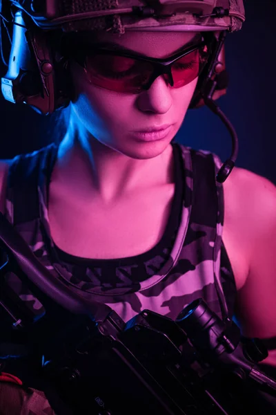 Flickan i militära overaller airsoft poserar med en pistol i händerna på en mörk bakgrund i dimman i neon ljus — Stockfoto