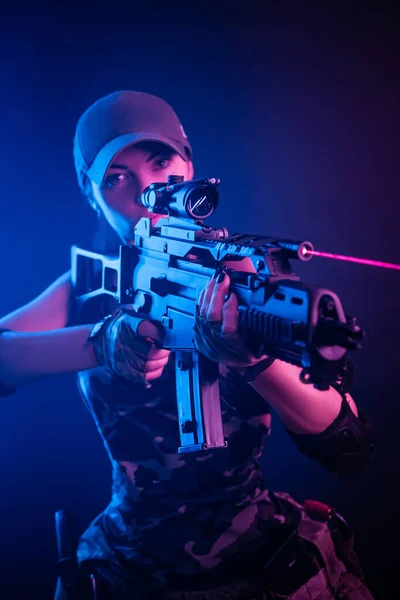 Askeri üniformalı kız, elinde silahla hava yumuşaklığında poz veriyor. Karanlık bir arka planda, neon ışıklı. — Stok fotoğraf