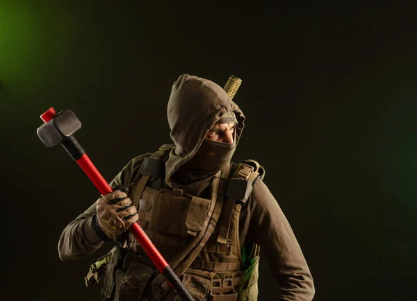 A soldier-saboteur rebel in military clothing with a weapon on a dark background holding a sledgehammer — Stock Fotó