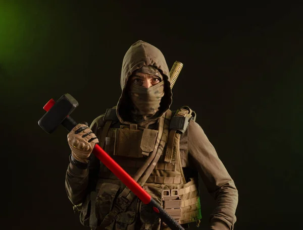 Um soldado-sabotador rebelde em roupas militares com uma arma em um fundo escuro segurando uma marreta — Fotografia de Stock