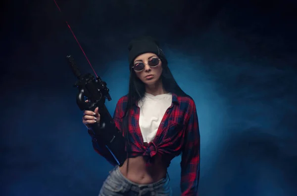 Una chica con camisa a cuadros sobre un fondo oscuro con un rifle automático — Foto de Stock