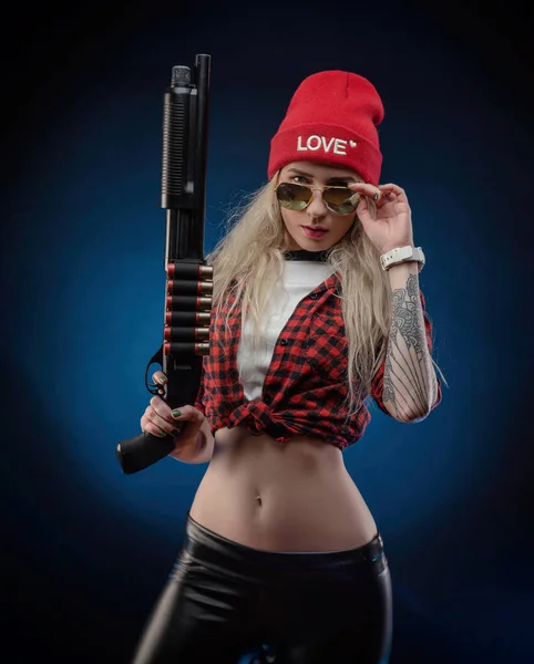 Una bionda luminosa in un cappello rosso con un fucile su uno sfondo scuro — Foto Stock