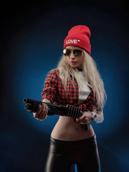 Une blonde lumineuse dans un chapeau rouge avec un fusil de chasse sur un fond sombre — Photo