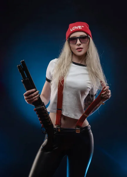 A bright blonde in a red hat with a shotgun on a dark background — Stock Photo, Image
