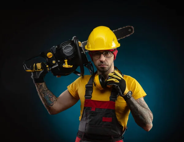 Een man in beschermende overalls met een kettingzaag op een donkere achtergrond — Stockfoto