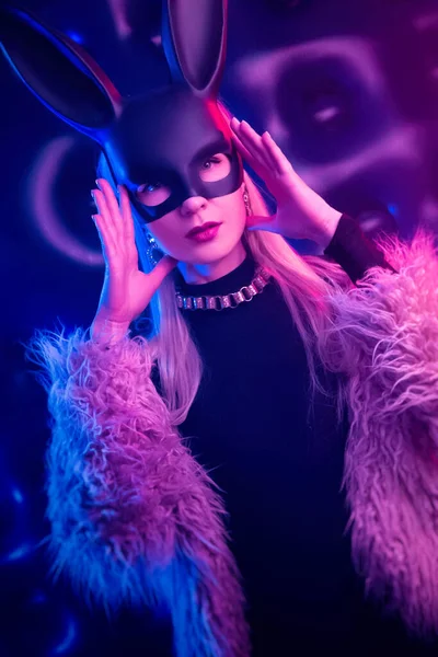 穿着紧身衣和皮毛外套，戴着兔子面具的性感女孩，在霓虹灯下 图库图片