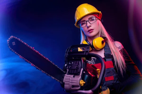 Een meisje met een kettingzaag in overalls op een zwarte achtergrond in een neon licht — Stockfoto