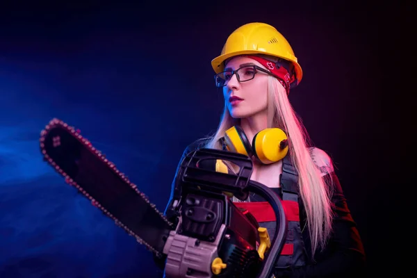 Uma menina com uma motosserra em macacão em um fundo preto em uma luz de néon — Fotografia de Stock