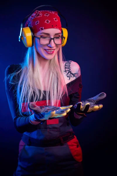 Una chica en overoles de construcción y auriculares sobre un fondo negro en luz de neón haciendo reparaciones de la casa — Foto de Stock