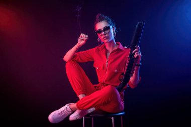 Parlak esmer, kırmızı tulumlu, siyah arka planda bir av tüfeği var.