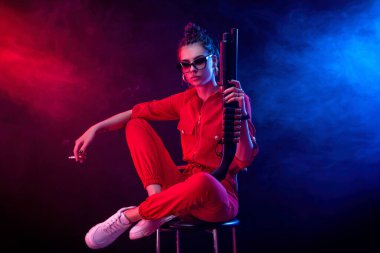 Parlak esmer, kırmızı tulumlu, siyah arka planda bir av tüfeği var.