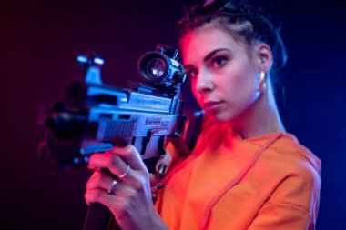 Kırmızı tulumlu güzel bir esmer, kısa namlulu bir makineli tüfek ve neon ışıklı bir arka planı var.