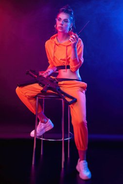 Parlak esmer, turuncu tulumlu ve arka planda makineli tüfekli, neon ışıklı. Radyoda konuşuyor.