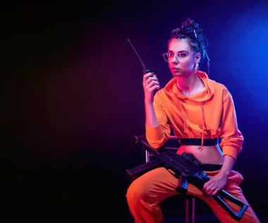 Parlak esmer, turuncu tulumlu ve arka planda makineli tüfekli, neon ışıklı. Radyoda konuşuyor.