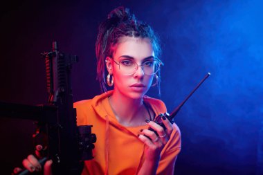 Parlak esmer, turuncu tulumlu ve arka planda makineli tüfekli, neon ışıklı. Radyoda konuşuyor.