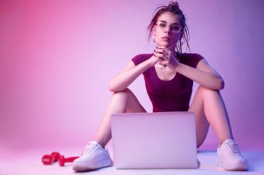 Cılız bir kız mayo giymiş, beyaz bir arka planda, neon ışıkta yerde oturuyor. Bilgisayarda çevrimiçi eğitim