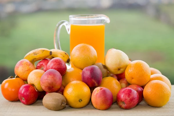 Szklanki soku pomarańczowego i mnóstwo owoców na drewnianym stole odkryty — Zdjęcie stockowe