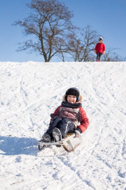 Güneşli bir kış gününde çocuklar tepeden aşağı kayıyor.