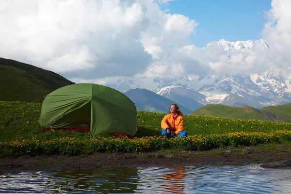 Jovem sentada perto de uma tenda em frente a picos de montanha nevados Fotografias De Stock Royalty-Free