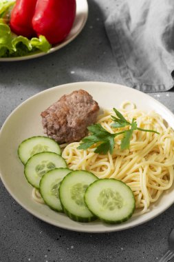 Gri bir masada bej bir tabakta makarna, pirzola ve salatalık.