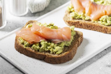 Açık gri mutfak masasında beyaz bir seramik tabakta avokado ve somon balığı. Avokado ve kırmızı balıklı kızarmış ekmek. Konsept yararlı ve lezzetli bir kahvaltı.