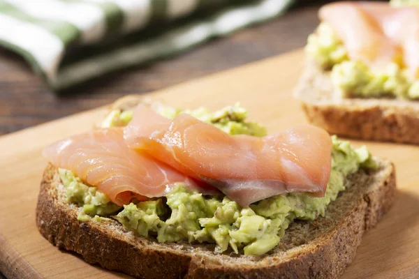Kahverengi ahşap bir masanın üzerinde, ahşap bir tahtanın üzerinde avokado ve somon balığıyla Avokado ve kırmızı balıklı kızarmış ekmek. Konsept kullanışlı ve lezzetli Kahvaltı