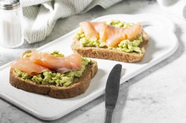 Açık gri mutfak masasında beyaz bir seramik tabakta avokado ve somon balığı. Avokado ve kırmızı balıklı kızarmış ekmek. Konsept yararlı ve lezzetli bir kahvaltı.