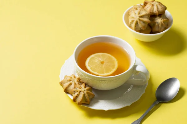 Tea with lemon in a white Cup and lemon cookies with poppy seeds in a white ceramic bowl on a yellow background. The concept of Breakfast or snack