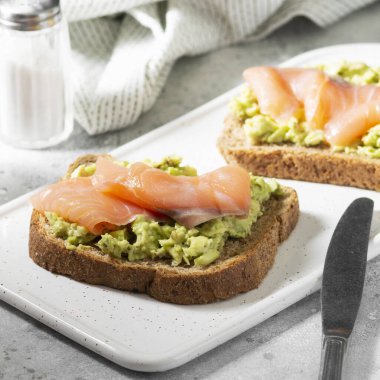 Açık gri mutfak masasında beyaz bir seramik tabakta avokado ve somon balığı. Avokado ve kırmızı balıklı kızarmış ekmek. Konsept yararlı ve lezzetli bir kahvaltı.
