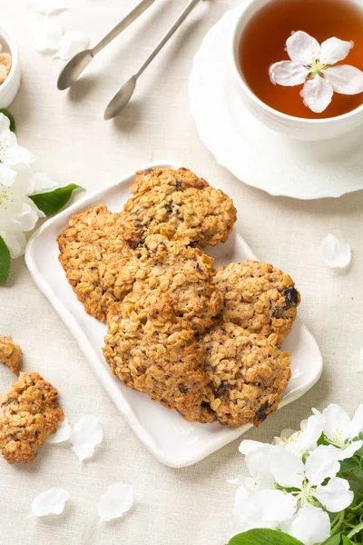 Oatmeal cookies cookies in a white ceramic plate on a light table among flowering branches. Healthy homemade oatmeal cookies. A plate of cookies and a Cup of tea on the table. The view from the top
