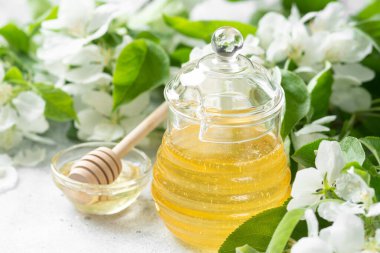 Çiçekli dalların arka planında cam bir kavanozda çiçek balı. Açık gri bir masada cam bir kavanozda sıvı bal. Bir kavanoz bal. Yaklaş.