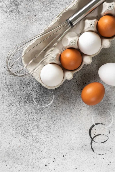 Eggs in a tray on the kitchen table. White and brown eggs with a whisk in the tray. Chicken eggs and a whisk on a culinary background. Concept of preparation for baking. Top view with space for text