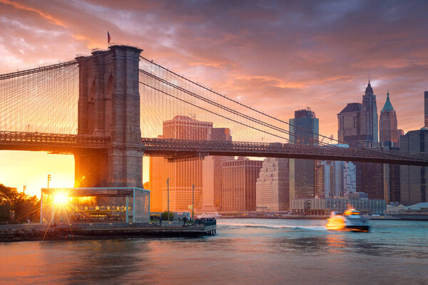 Famous Brooklyn Bridge in New York City with financial district - downtown Manhattan in background. Sightseeing boat on the East River and beautiful sunset over Jane's Carousel.