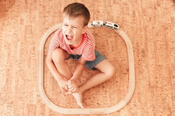 Förtjusande pojke som sitter på golvet med träjärnväg runt sig — Stockfoto