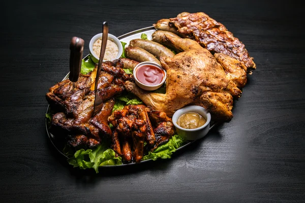 Grilled meat, vegetables, chicken, hot dogs, sausages grilling, sauce, salad served on a plate on a black background