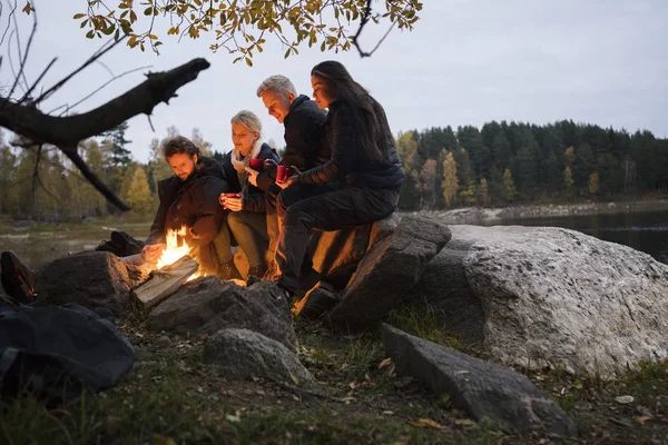 Jóvenes amigos que se relajan junto a la fogata en la orilla del lago —  Fotos de Stock