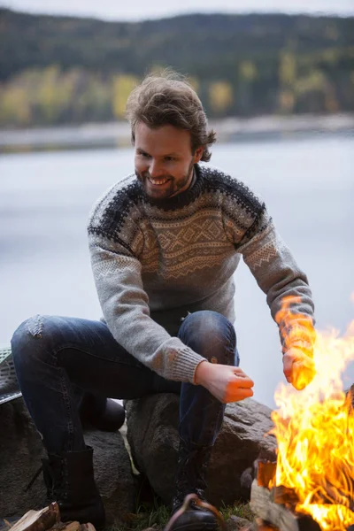 Hombre calentando manos por hoguera en camping junto al lago — Foto de Stock