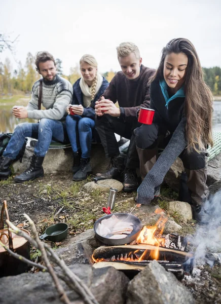 Mujer cocina comida en fogata con amigos en segundo plano — Foto de Stock