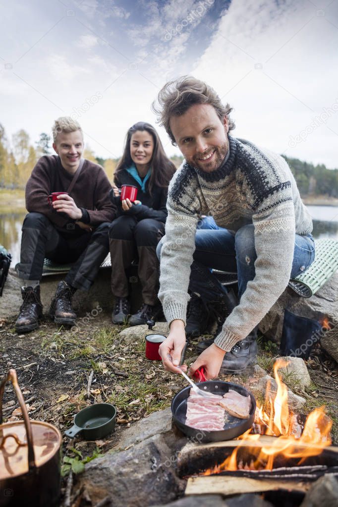 Man Cooking Food On Campfire With Friends In Background