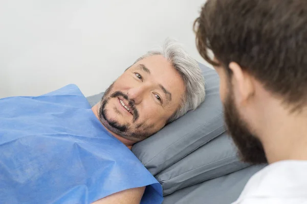 Paciente do sexo masculino deitado na cama do hospital enquanto olha para o médico — Fotografia de Stock