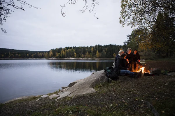 Unga vänner sitter nära brasa vid sjön i skymningen — Stockfoto