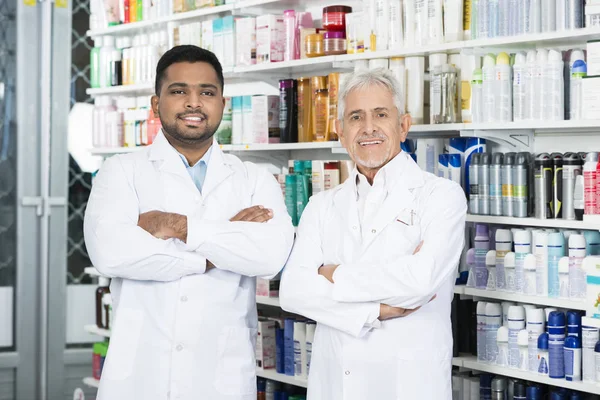 Braços permanentes multiétnicos do farmacêutico cruzados na farmácia — Fotografia de Stock