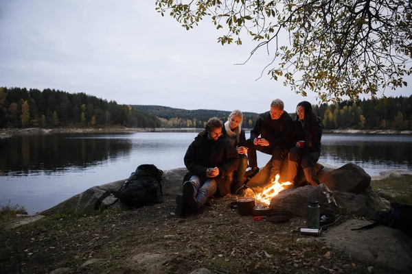 Unga vänner med kaffekoppar som sitter nära lägerelden — Stockfoto