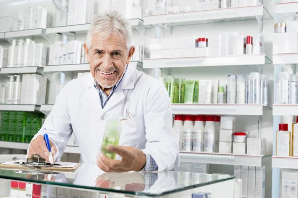 Sênior farmacêutico segurando produto enquanto escrevendo na área de transferência — Fotografia de Stock