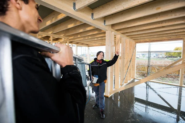 Carpintero haciendo gestos mientras su colega lleva escalera en la construcción — Foto de Stock
